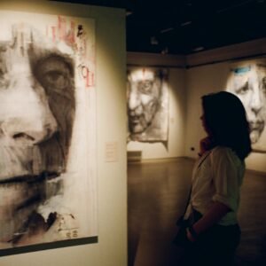 A woman observes portrait paintings in a dimly lit modern art gallery.